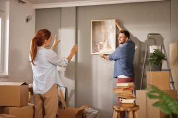 A couple is in the process of decorating their new home as one partner hangs a framed picture on...