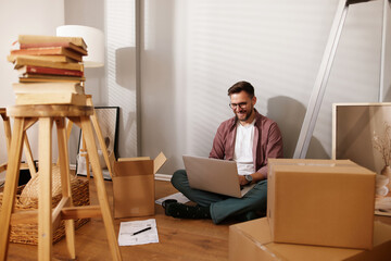 Individual sits on the floor, engaging with a laptop amidst unpacked moving boxes and stacks of...