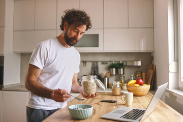 A man is preparing oatmeal for breakfast in a stylish kitchen. He holds a jar of oats and looks...