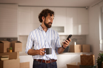 A man stands in his new home, smiling as he looks at his phone while holding a coffee cup. Moving...