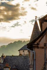 The glow of sunset on ancient stone rooftops with a bird weather vane on top of the bell tower in the traditional French village of Conques, France, under a dramatic sky backlit by the setting sun.