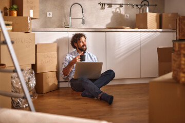 A man sitting on the floor surrounded by unpacked boxes in his new kitchen, sipping a drink while...