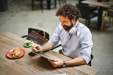 A man engages with a tablet, focused on his work, while seated at a wooden table. He enjoys a cup of coffee and a pastry, with headphones around his neck, in a contemporary workspace.