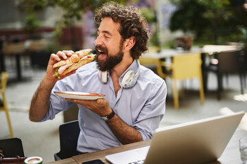 A man with headphones takes a bite of a sandwich while sitting at an outdoor table, enjoying a...