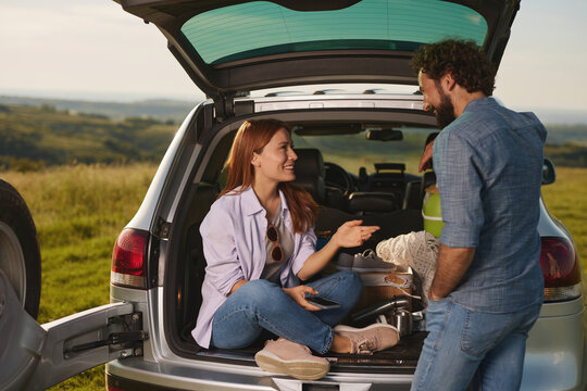 Friends gather near their vehicle for a cozy picnic, sharing food and laughter while seated comfortably in the open trunk, surrounded by scenic green hills. - Powered by Adobe