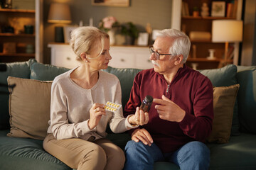 An elderly man and woman sit on a teal sofa, discussing medication. The man points to a bottle, while the woman holds pills.