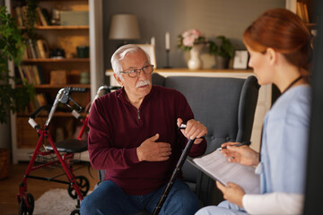 An elderly man with a mustache sits in a gray armchair holding a cane, talking to a red-haired...