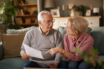 An elderly couple sits closely on a blue couch, reviewing financial documents. The man points to a document while speaking to the woman. Books and decorations adorn the background.