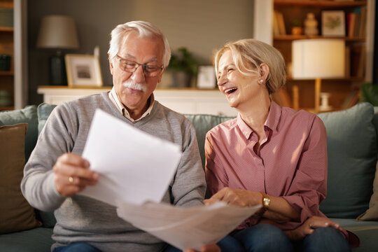 A senior man with glasses reads papers while a smiling senior woman sits beside him on the sofa in their home. They appear to be reviewing financial documents in the afternoon.