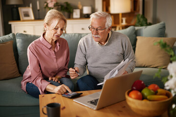 Two senior adults, a man and a woman, sit on a couch at home. They review paperwork and use a...