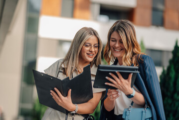 Businesswomen smiling and comparing notes on tablet and clipboard outdoors