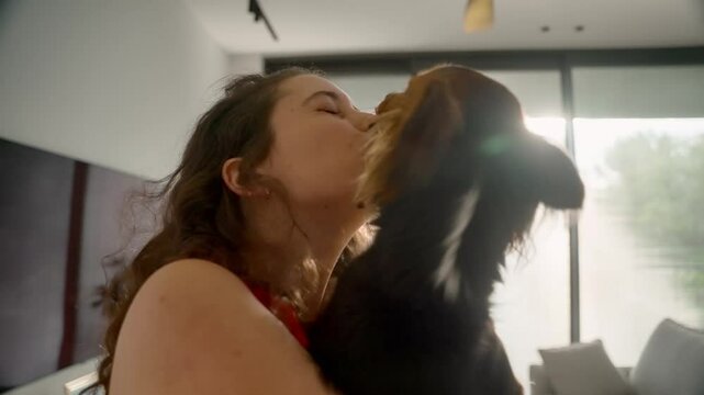 Female pet owner and her cute little dog hugging and enjoying moment together. Small furry canine licking young woman in joy as she is laughing and dancing having fun in apartment living room