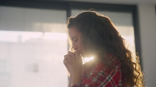 Smiling and cheerful young woman is praying with sun shining behind her. Hands clasped or folded together she is making a wish asking something or having private moment or prayer and religion indoors