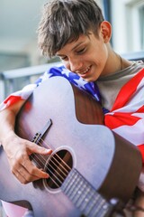 Happy teenager with American flag on shoulders playing guitar on balcony