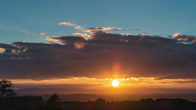 Breathtaking sunrise over a forested landscape with the sun peeking through dramatic clouds. Soft golden light spreading across the horizon, highlighting trees and hills in the early morning