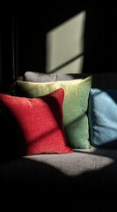 Sunlit velvet pillows in red, green, and blue hues rest on a dark gray sofa, dramatic shadows accentuate the textures