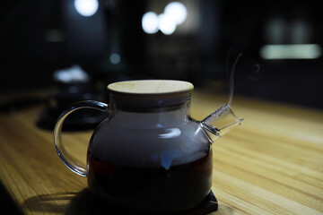 Glass Teapot with Wooden Lid and Steam on Wooden Table in Dim Lighting