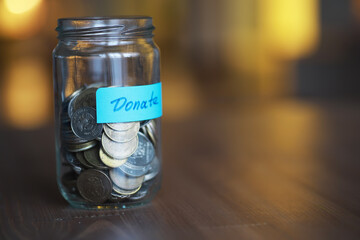 Glass Jar Filled with Coins for Donation on Brown Wooden Table with Warm Yellow Light Background