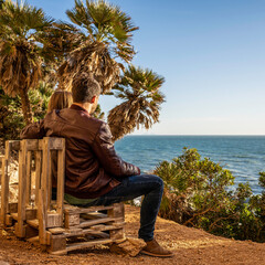Couple sitting on a rustic bench made of wooden pallets enjoying the ocean view under palm trees. Warm afternoon light and peaceful atmosphere. Romantic moment and nature lifestyle concept.