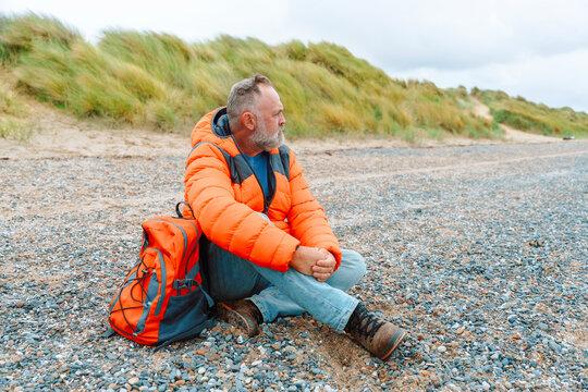 Man in orange jacket sitting on rocky beach with backpack during cloudy afternoon near grassy dunes