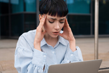 Young woman experiencing stress from work while sitting outdoors with a laptop in a modern urban...