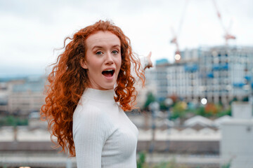 Young woman with curly red hair smiles enthusiastically against a cityscape backdrop during an...