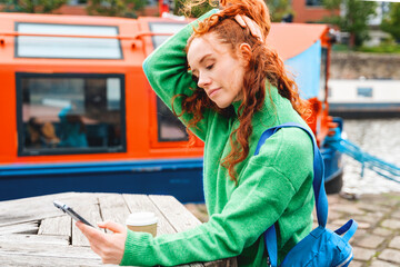 Woman in green sweater checks her phone while sipping coffee near a colorful boat by the water...