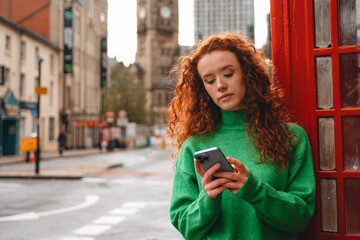 Young woman with curly hair in green sweater checks her phone near a traditional telephone booth on...