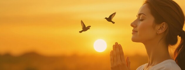 Woman meditating with hands in prayer pose against a golden sunset sky with birds flying, symbolizing peace and spirituality.