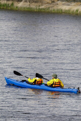 Two people in yellow jackets and life vests paddle a blue tandem kayak across calm lake waters...
