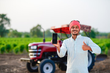 young indian farmer standing with tractor and showing smartphone at field
