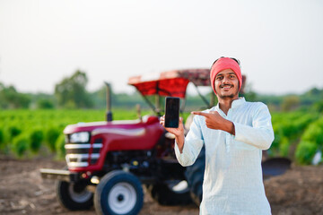 young indian farmer standing with tractor and showing smartphone at field
