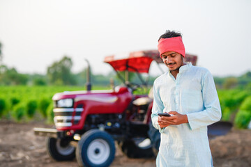 indian farmer standing with tractor and talking on smartphone at field