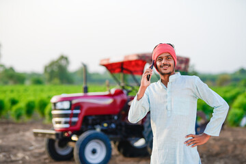 indian farmer standing with tractor and talking on smartphone at field