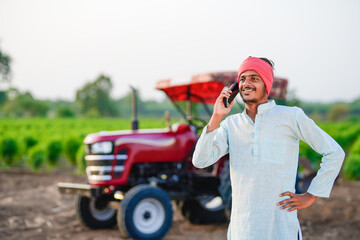 indian farmer standing with tractor and talking on smartphone at field