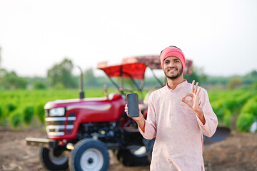young indian farmer standing with tractor and showing smartphone at field