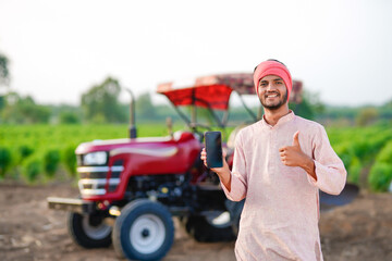 young indian farmer standing with tractor and showing smartphone at field