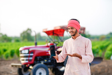 young indian farmer standing with tractor and showing smartphone at field