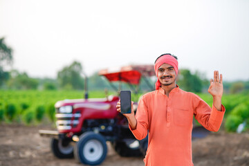 young indian farmer standing with tractor and showing smartphone at field