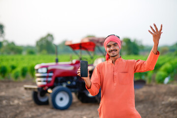 young indian farmer standing with tractor and showing smartphone at field