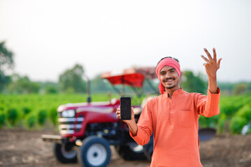 young indian farmer standing with tractor and showing smartphone at field