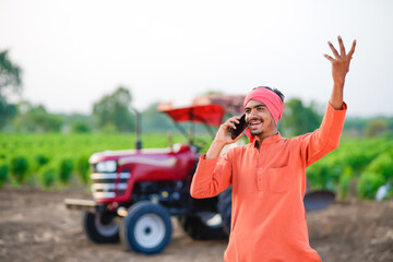 indian farmer standing with tractor and talking on smartphone at field