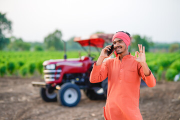 indian farmer standing with tractor and talking on smartphone at field