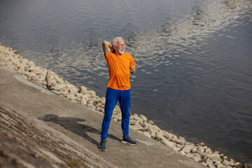 Full length of senior sportsman in shape standing on riverbank and stretching his arms.