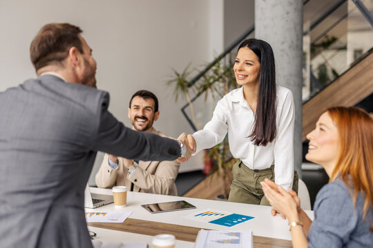 Smiling businesswoman standing at corporate office with her manager and shaking hands while other team members applauding.