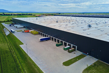 Large modern warehouse with loading docks and parked trucks surrounded by green fields. Industrial...