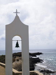 White bell tower with a single bell, framed by rocky coastline and cloudy sky in the background.