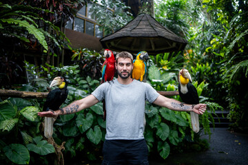 Man interacting with colorful exotic birds, including macaws and hornbills, in a tropical jungle setting. Concept of adventure, wildlife tourism, biodiversity and unique travel experiences.

