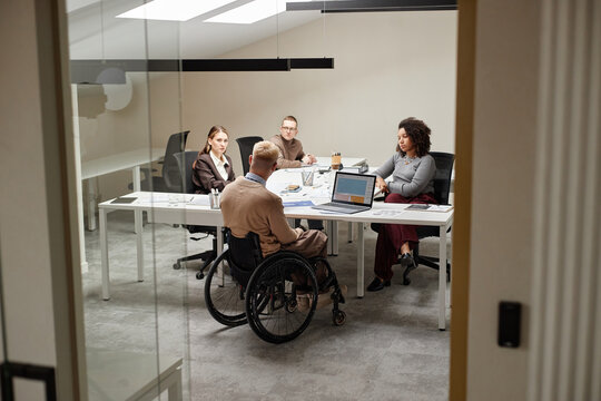 Diverse group of young adult professionals including Caucasian man with disability in wheelchair discussing business strategy around conference table in modern office