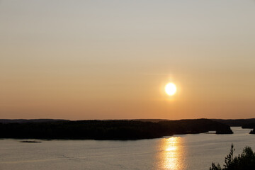 The sun sets over a calm forest lake, casting golden reflections on the water under a warm, clear sky.